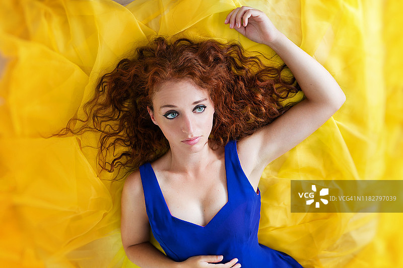 身穿蓝色无袖连衣裙的红发女郎躺在薄薄的黄色织物上，凝视着远方图片素材