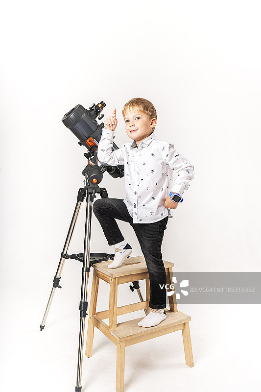 一个戴眼镜的小男孩站在椅子上的大反射望远镜前。图片素材