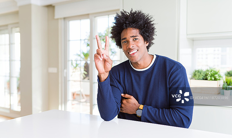 年轻的非洲裔美国人穿着休闲毛衣坐在家里微笑着看着摄像机显示手指做胜利手势。二号人物。图片素材