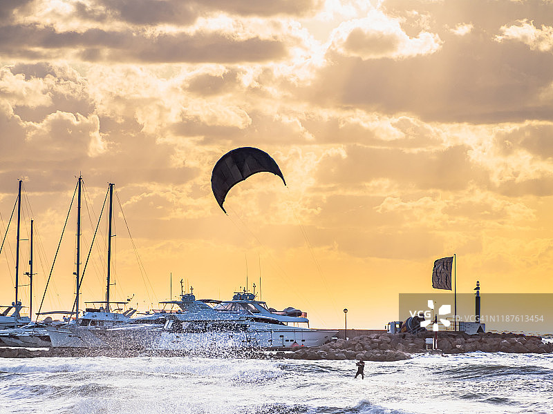 日落时分，一个人在海上练习风筝冲浪，巨大的海浪拍打着港口的船只和游艇图片素材