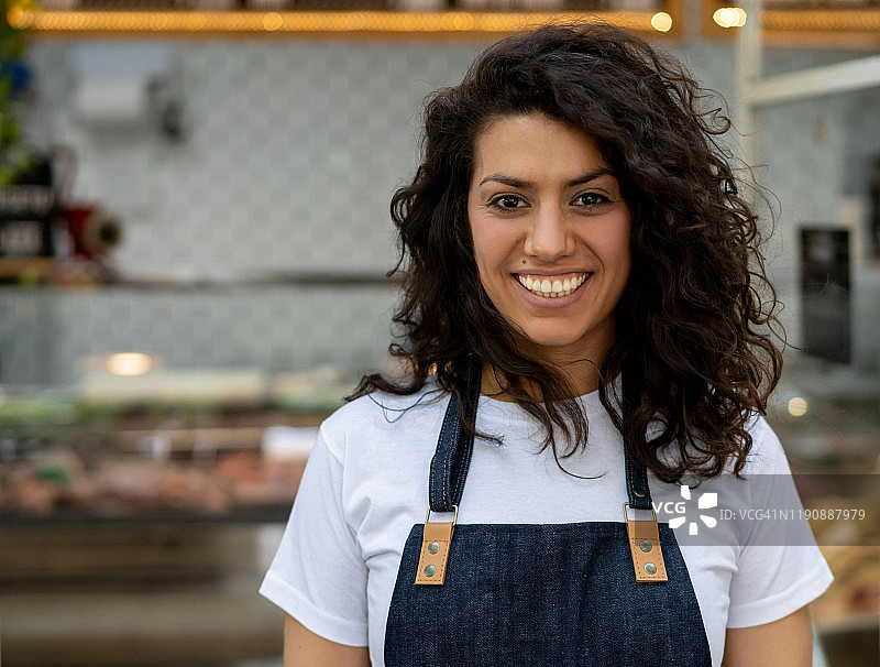 拉丁美洲美丽的女雇员在肉店穿着围裙对着镜头微笑图片素材