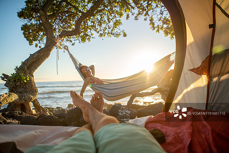 男人的脚从一个帐篷露营在夏威夷的海滩看女朋友在吊床户外的观点图片素材