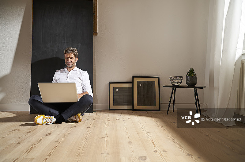 放松成熟的男人坐在家里的地板上使用笔记本电脑图片素材