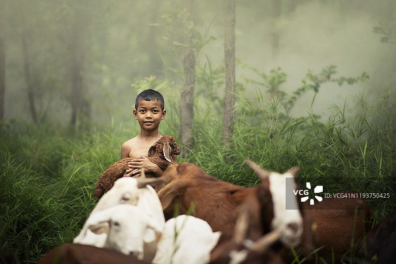 在一片绿色的森林里，一个微笑的男孩抱着一只小山羊图片素材