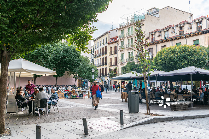 在马德里的Malasana社区，一名妇女经过一个广场，那里的人们正在户外用餐。图片素材
