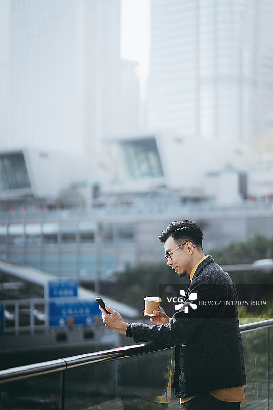 自信的画像，年轻的亚洲商人用智能手机，喝着咖啡，在现代写字楼前与城市风景相反图片素材