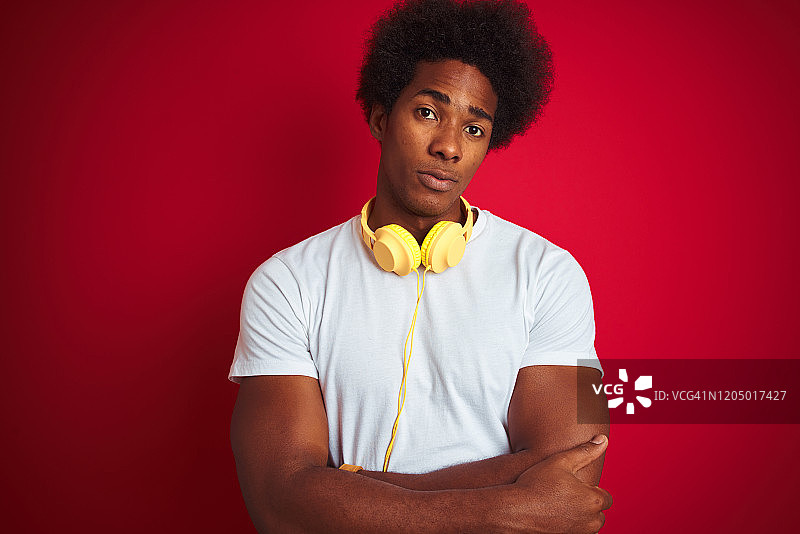 年轻的非洲裔美国人听音乐使用耳机在孤立的红色背景怀疑和紧张，不赞成的表情在脸上交叉双臂。消极的人。图片素材