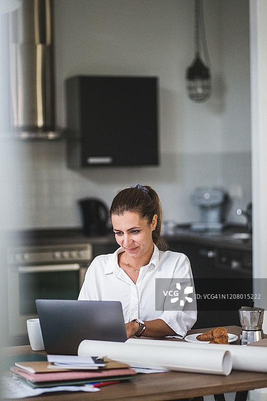 微笑的女性自由职业者使用笔记本电脑，坐在家里的桌子旁图片素材