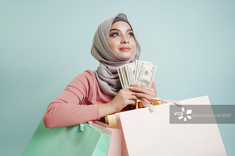 快乐的亚洲穆斯林妇女手里拿着美元和购物袋正在考虑要买的东西。富家女，金融投资概念。图片素材