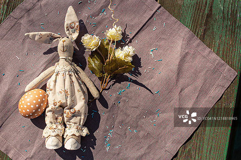 手捧鲜花的手捧玩具兔兔和绘有各种白色图案的棕色彩蛋庆祝复活节。前视图。带有阳光的紫色棉布桌布。东欧正统文化图片素材