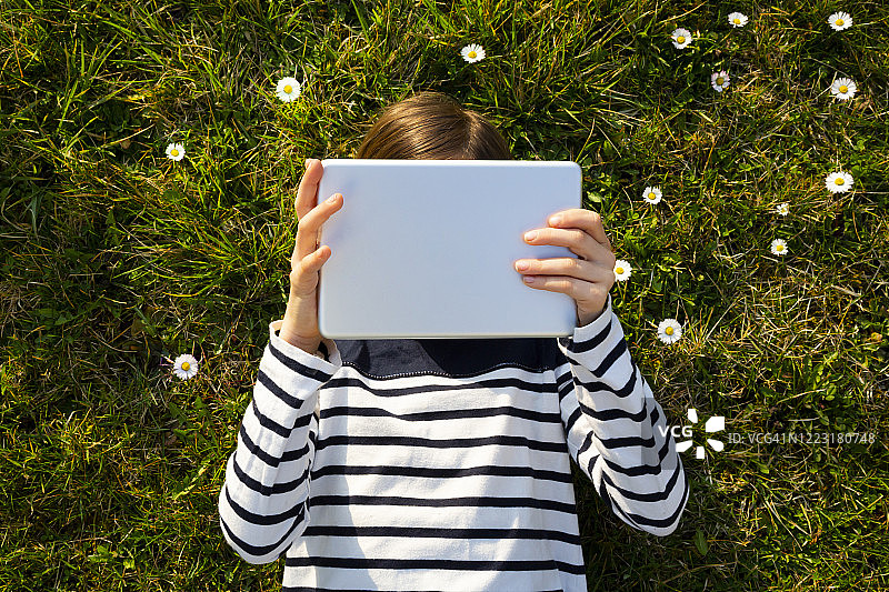 小女孩躺在鲜花草地上，把脸藏在数码平板电脑后面图片素材
