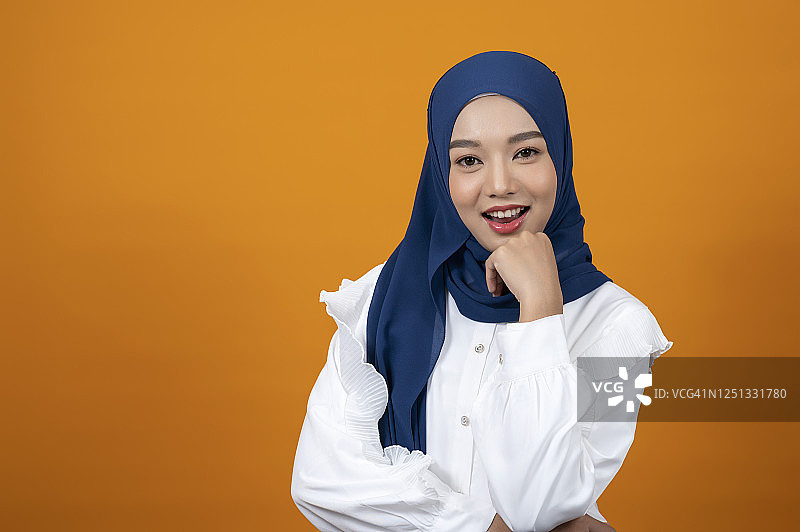 摄影棚拍摄了一名穆斯林妇女微笑着用手托着下巴，戴着头巾站在浅橙色的背景前。图片素材