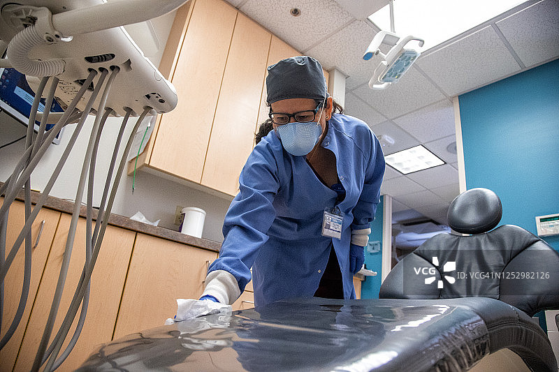 一名50多岁的拉丁裔保健工作者戴着口罩和手术手套在牙科诊所的检查室擦拭医疗椅，为下一个病人做准备图片素材