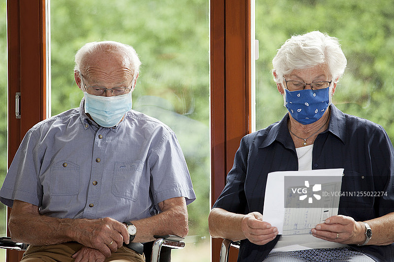 一对戴着口罩的老年夫妇坐在医院或办公室明亮的候诊室里图片素材