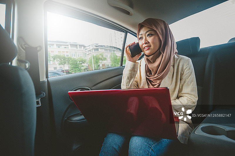 年轻的亚裔穆斯林女企业家坐在车里，拿着笔记本电脑，在打电话图片素材