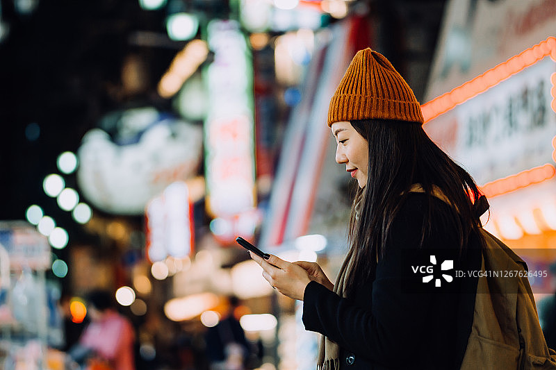 时尚的年轻亚洲女性旅行者背包使用智能手机上的移动应用设备导航，在繁忙的市中心街道上探索夜晚图片素材