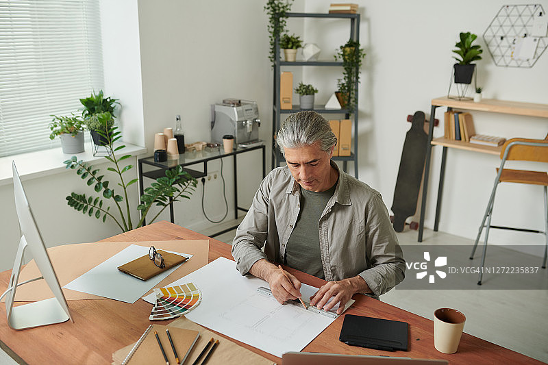 集中成熟的白发男子坐在家里办公室的办公桌上盆栽和工作与素描图片素材