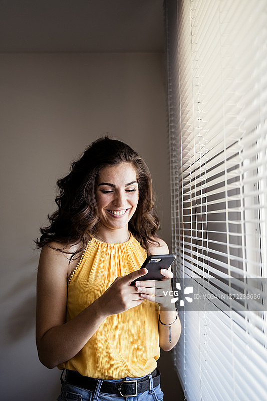 一名快乐的年轻女子站在家里的百叶窗旁，用智能手机发短信图片素材