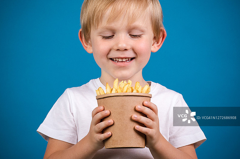 一个白发男孩的特写看着一个一次性纸杯与快餐薯条，并即将吃它。工作室用蓝色背景拍摄。图片素材