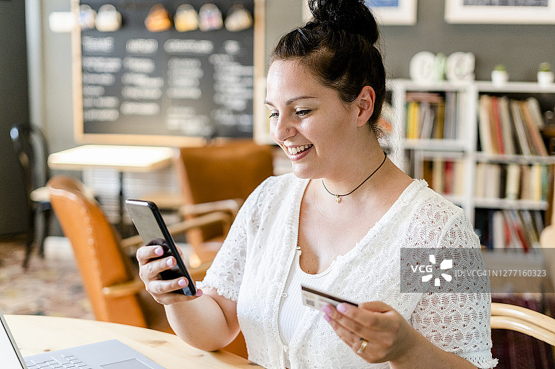 一个微笑的年轻女子在咖啡店用智能手机拿着信用卡的特写图片素材