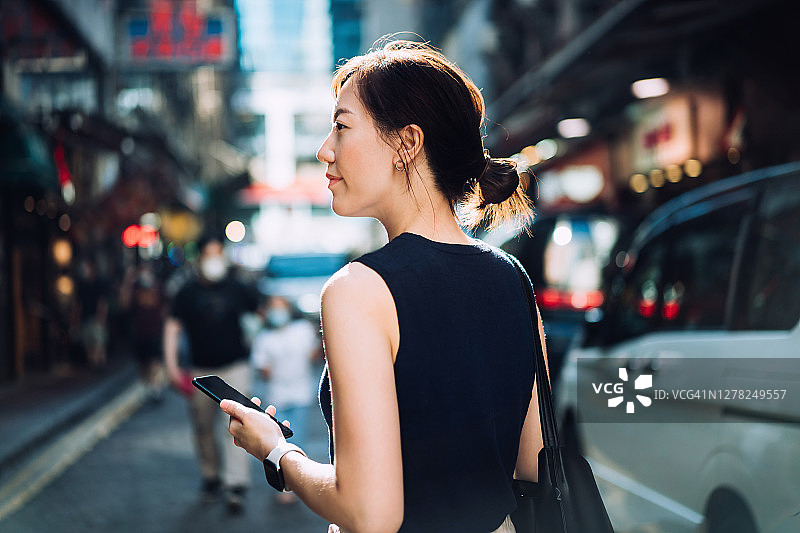 美丽的年轻亚洲女子手拿智能手机，在繁忙的市中心街道上与汽车和城市建筑擦肩而过图片素材