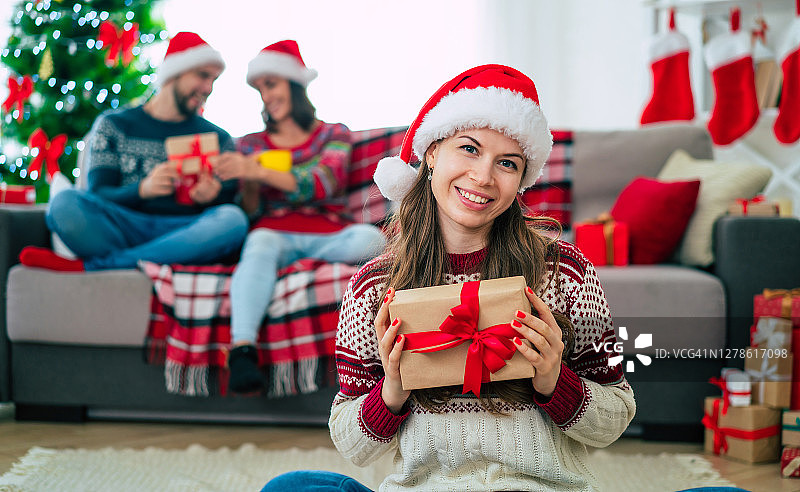 在她的朋友和圣诞树的背景上，美丽的年轻快乐微笑的女人穿着圣诞毛衣和圣诞帽，手里拿着一个彩色的礼物盒在家里。图片素材