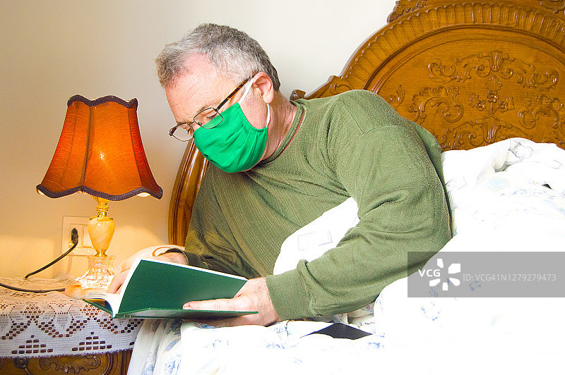 (Covid-19)男子躺在家里的床上，戴着绿色口罩看书图片素材