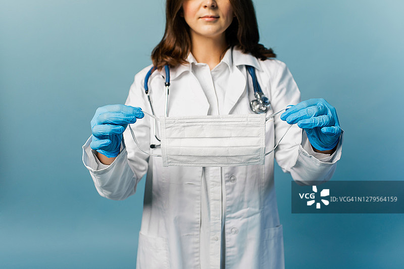 近近的照片女性医生在白色的实验室大衣拿着白色面具在蓝色的手套，而站在蓝色的背景。图片素材