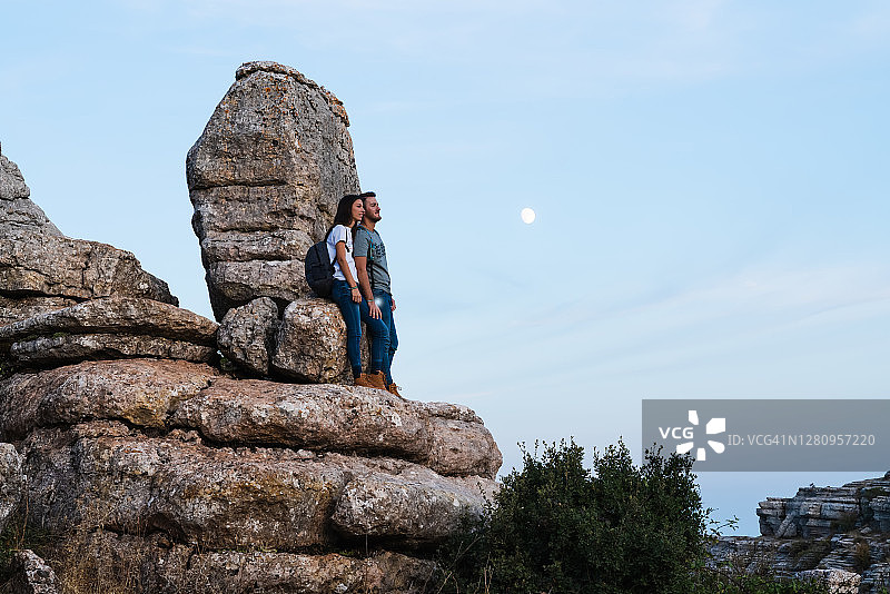 一对年轻的夫妇站在一个巨大的岩层上看着。到月亮旁边的地平线图片素材
