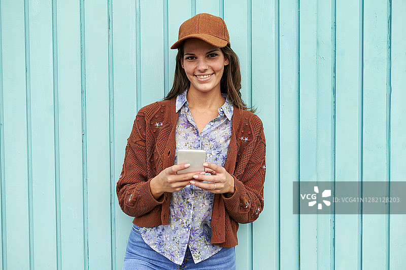 面带微笑的女人站在蓝色的金属门前，用手机发短信图片素材