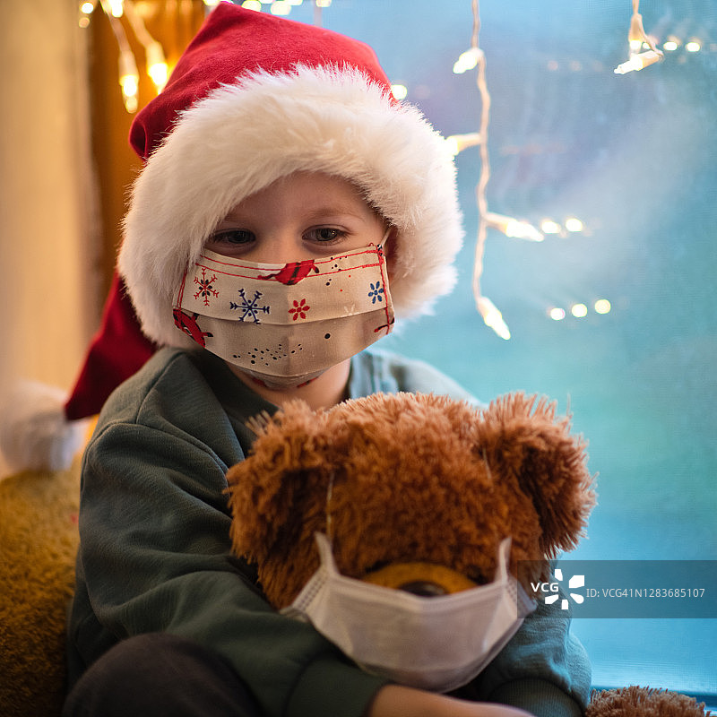 可爱的小男孩戴着防护面具和圣诞帽和他的泰迪熊在圣诞节图片素材