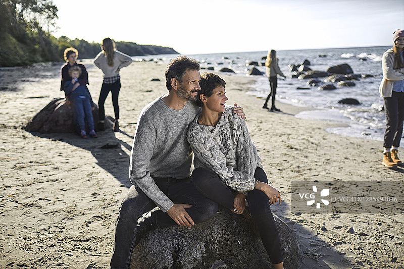 一名成年男子和一名年轻女子坐在沙滩上的巨石上，背景是家人图片素材