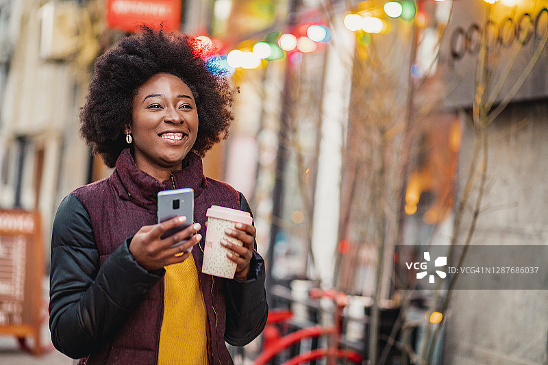 非裔美国妇女手持手机和可重复使用的咖啡杯走在街上的肖像图片素材