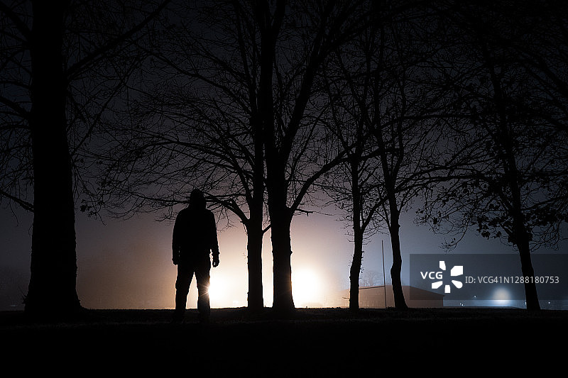 一个戴着兜帽的身影站在树旁，在明亮的灯光映衬下形成剪影。在一个有雾的冬天的晚上。图片素材