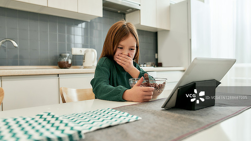 一个小女孩正在看动画片在她的平板电脑和咯咯笑，而独自坐在桌子上吃快速早餐在远程教育图片素材
