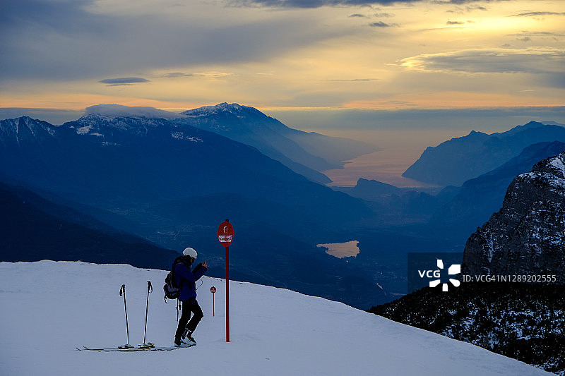 男孩戴着头盔，在夕阳下的滑雪坡上，光线反射在加尔达湖和阿尔卑斯山的雪峰上图片素材