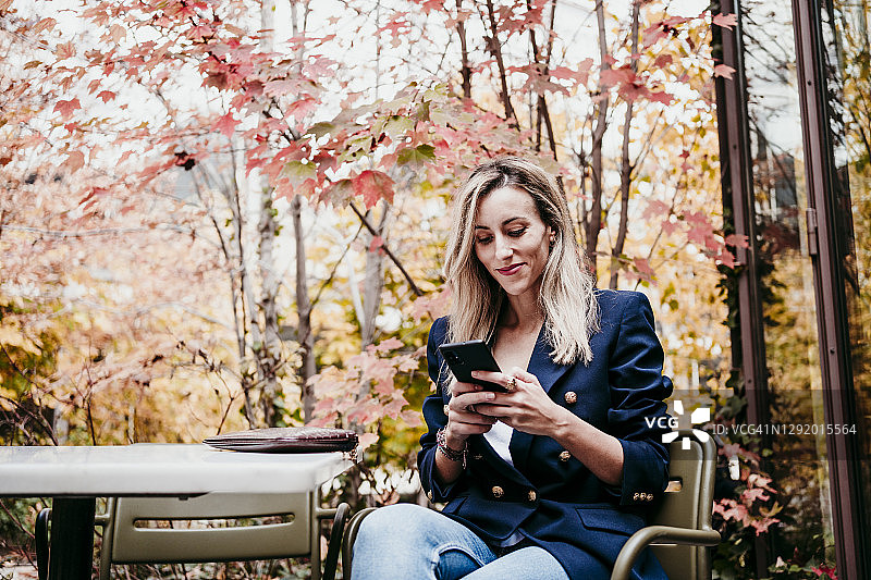 金发的中年女人坐在露台上对着秋树玩智能手机图片素材