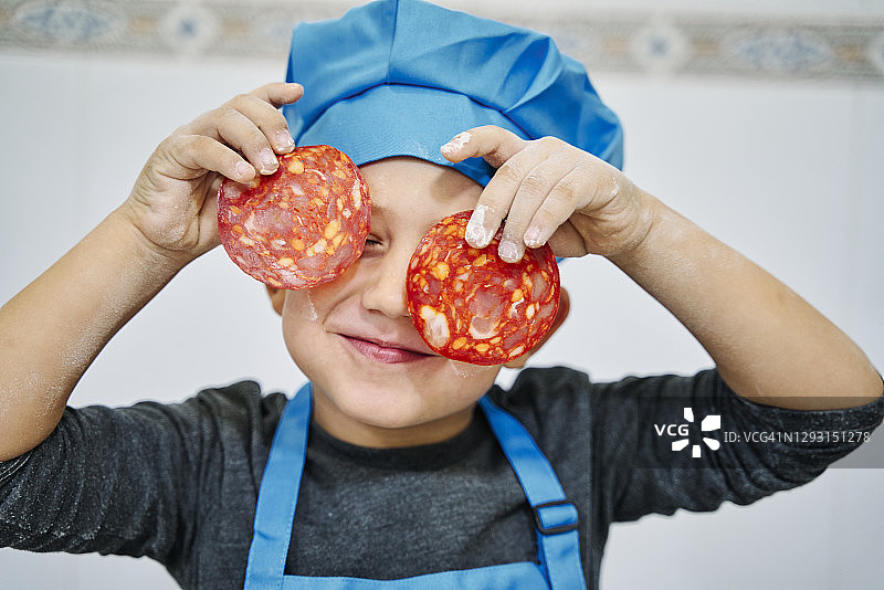 小男孩扮成厨师戴着蓝帽子拿着两片意大利香肠图片素材