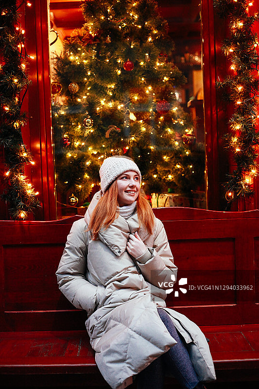 红头发的年轻快乐微笑的女人穿着温暖的冬装在街头，圣诞节日市场装饰着仙女灯和树在夜晚。生活方式度假。图片素材