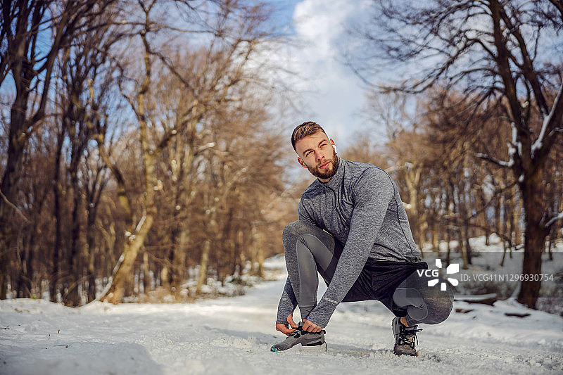 在冬天下雪的日子里，跑步者蹲在森林里的小路上系鞋带。健康的生活方式，运动服，下雪的天气图片素材