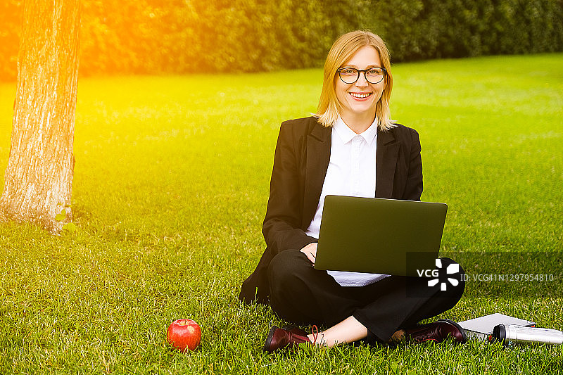 一个快乐美丽的女孩或女人戴着眼镜坐在草地上，用笔记本电脑工作，看着相机，微笑着。旁边有一本书或笔记本，一个热水瓶，一个苹果。这个学生在户外学习。远程教育的概念。图片素材