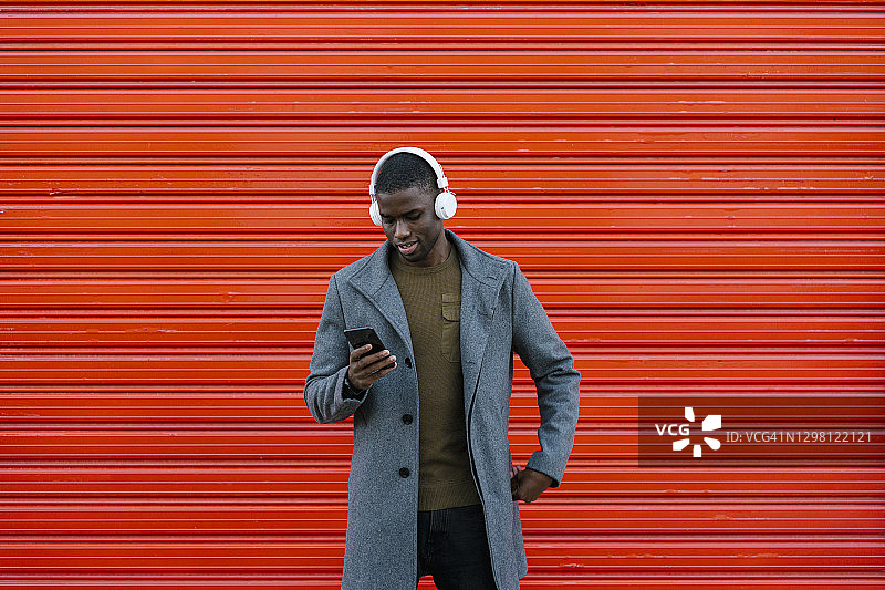 微笑的男人戴着耳机，用手机，而站在红色的墙图片素材