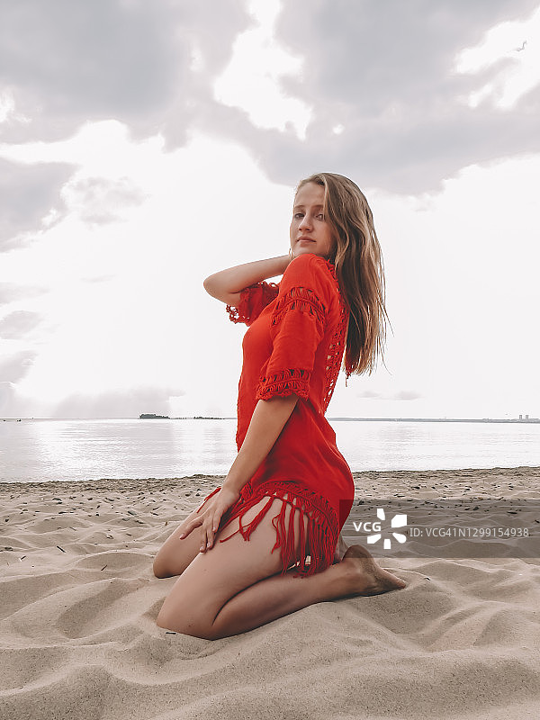 一位皮肤黝黑的年轻女子，身材苗条迷人，一头金色长发，身穿红色夏装，在沙滩上摆姿势，背景是海水和如画般的天空图片素材