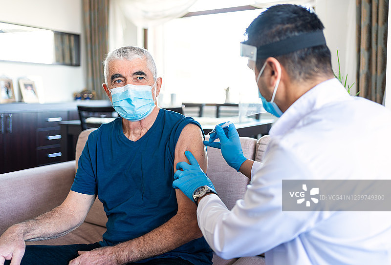 佩戴防护口罩的男护士在冠状病毒爆发期间坐在家中给一名老年男子注射。老年患者在家接种疫苗。冠状病毒疫苗接种图片素材