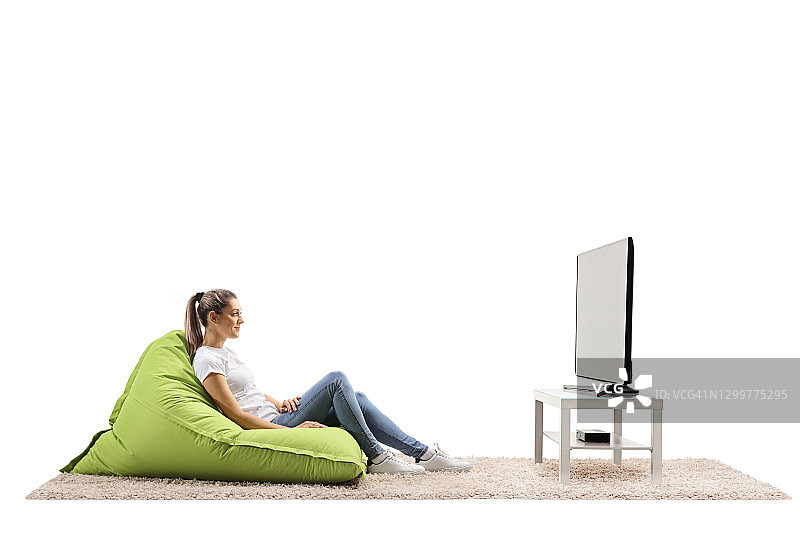 侧面拍摄的一个随意的年轻女子坐在绿色豆袋扶手椅和看电视图片素材