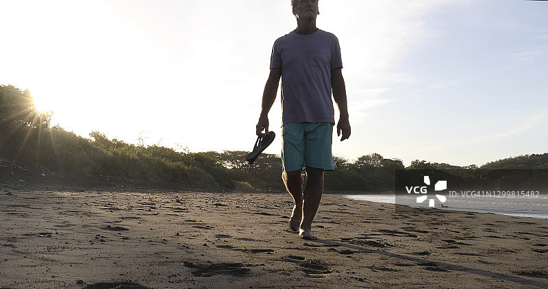 日出时分，一名男子手拿凉鞋走下沙滩图片素材