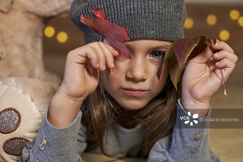 一个可爱的小女孩的肖像，有着大大的蓝眼睛和灰色的羊毛帽子躺在床上，玩红色的树秋天的叶子。图片素材