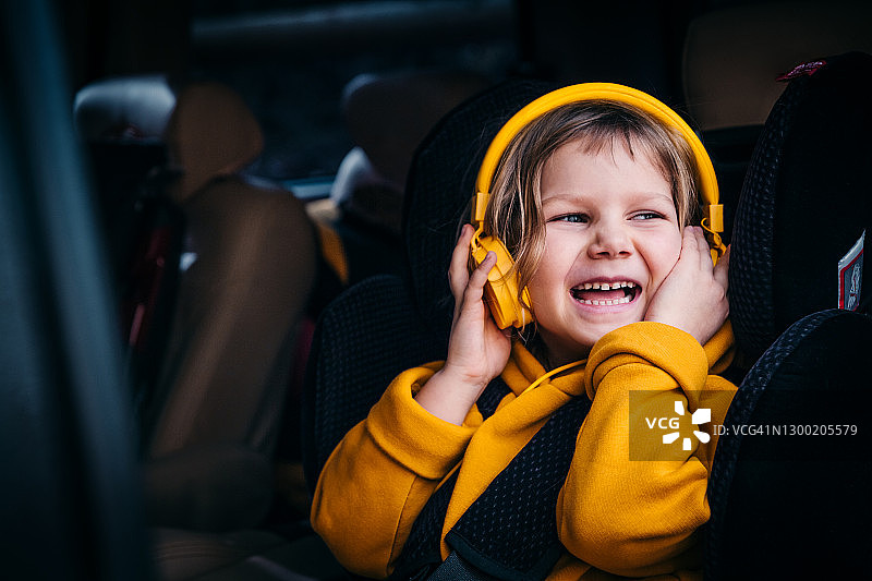 这孩子正坐在汽车座椅上。一个穿着黄色衣服的小女孩戴着耳机听着音乐，在车里玩得很开心。家庭旅游的概念。图片素材