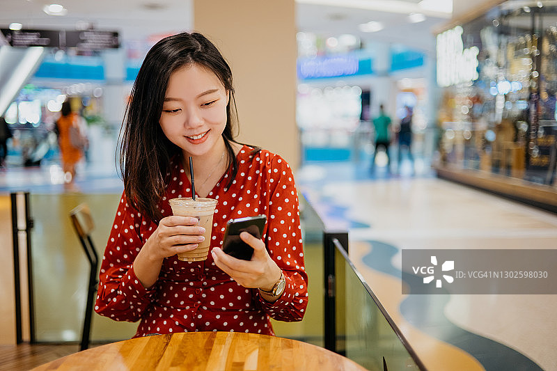 年轻的亚洲女性拿着咖啡杯，在咖啡馆里使用智能手机图片素材