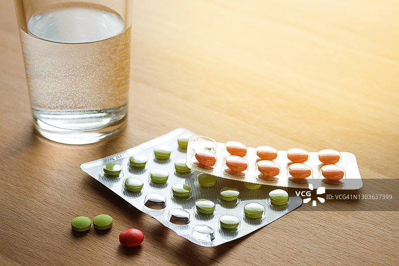 红色和绿色的药丸和一杯水放在厨房的木制桌子上。服用维生素、止痛药、药物和膳食补充剂。疾病的概念预防和治疗，保健和医药。计划和准备怀孕。图片素材